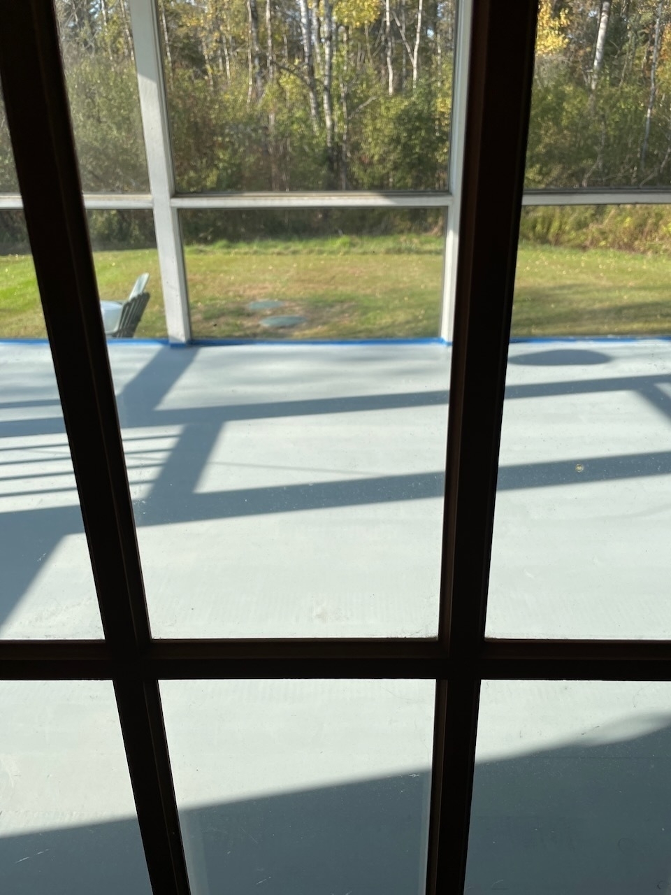 A view through a grid-patterned window shows a sunlit porch floor and a green lawn surrounded by trees.